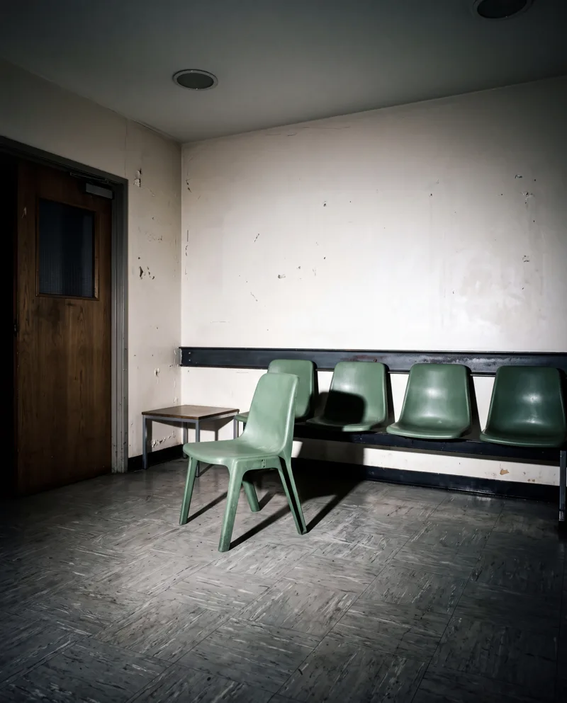 A waiting room after hours: a row of green plastic chairs against the wall and one pulled out of the line, empty.