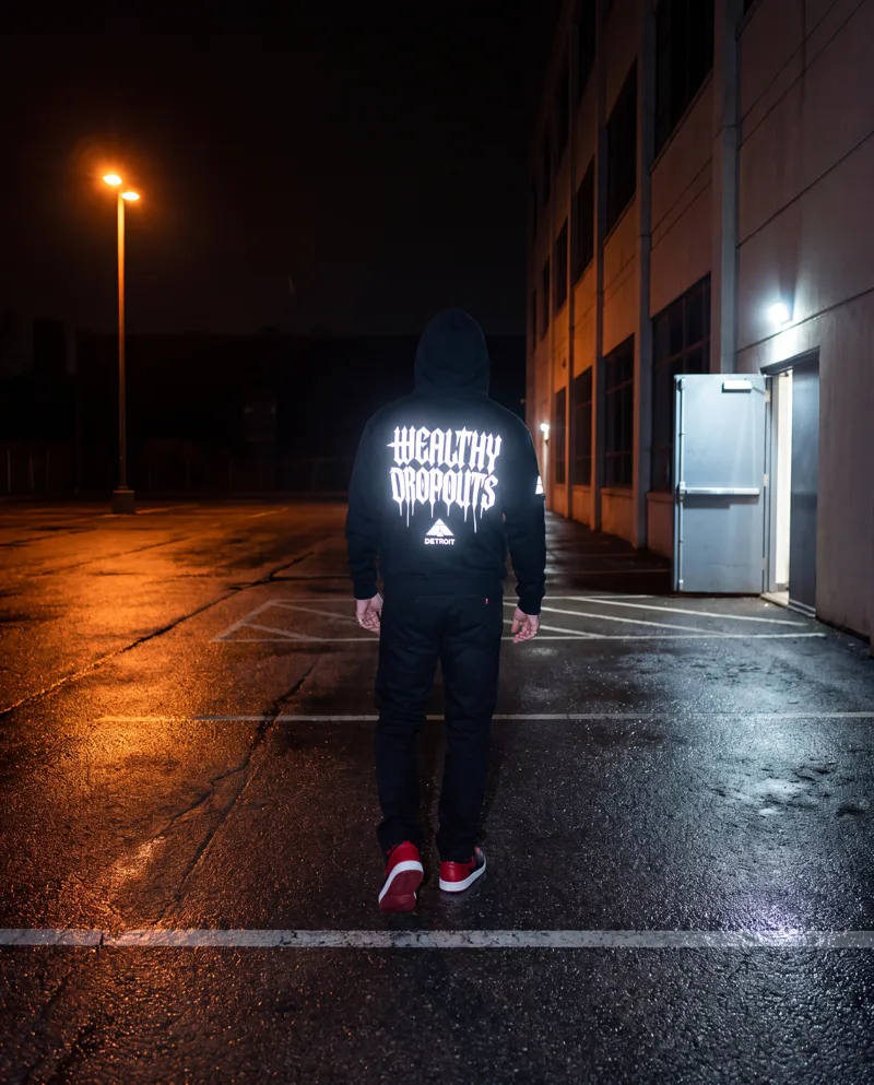 From behind: a man in a black hoodie walks away across a wet, empty lot at night under one lamp, WEALTHY DROPOUTS lit white across his back.