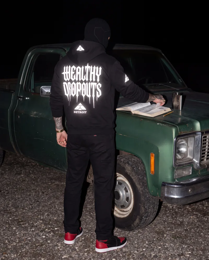 From behind: he stands at the bonnet of an old green pickup in the dark, turning a page of a workshop manual, WEALTHY DROPOUTS lit white across his back.