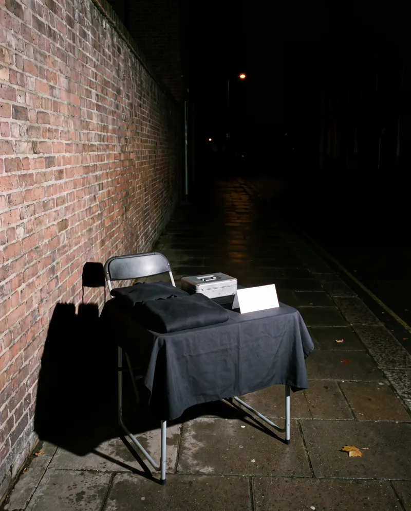 A folding table under a black cloth on an empty pavement at night: two folded black hoodies, a cash box and a blank card. Nobody is there.