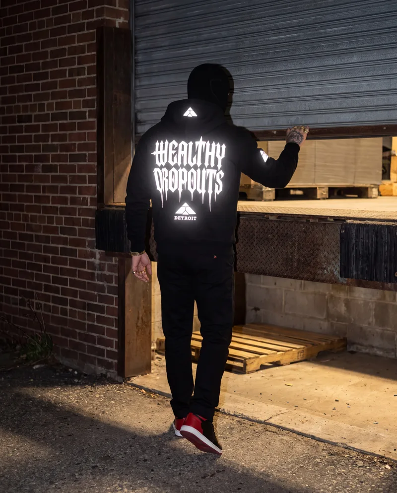 From behind: he lifts a half open loading door and steps toward the light under it, WEALTHY DROPOUTS lit white across his back.