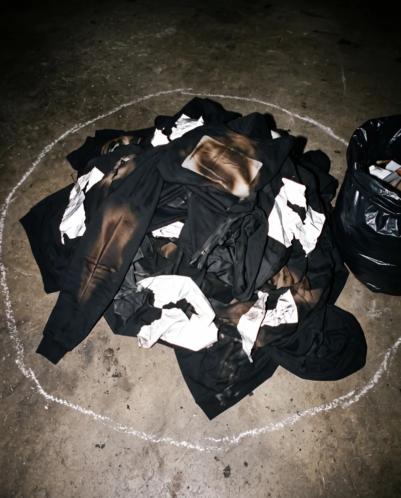 A pile of ruined black hoodies on a concrete floor, scorched, the silver film peeling, ringed in chalk like evidence.
