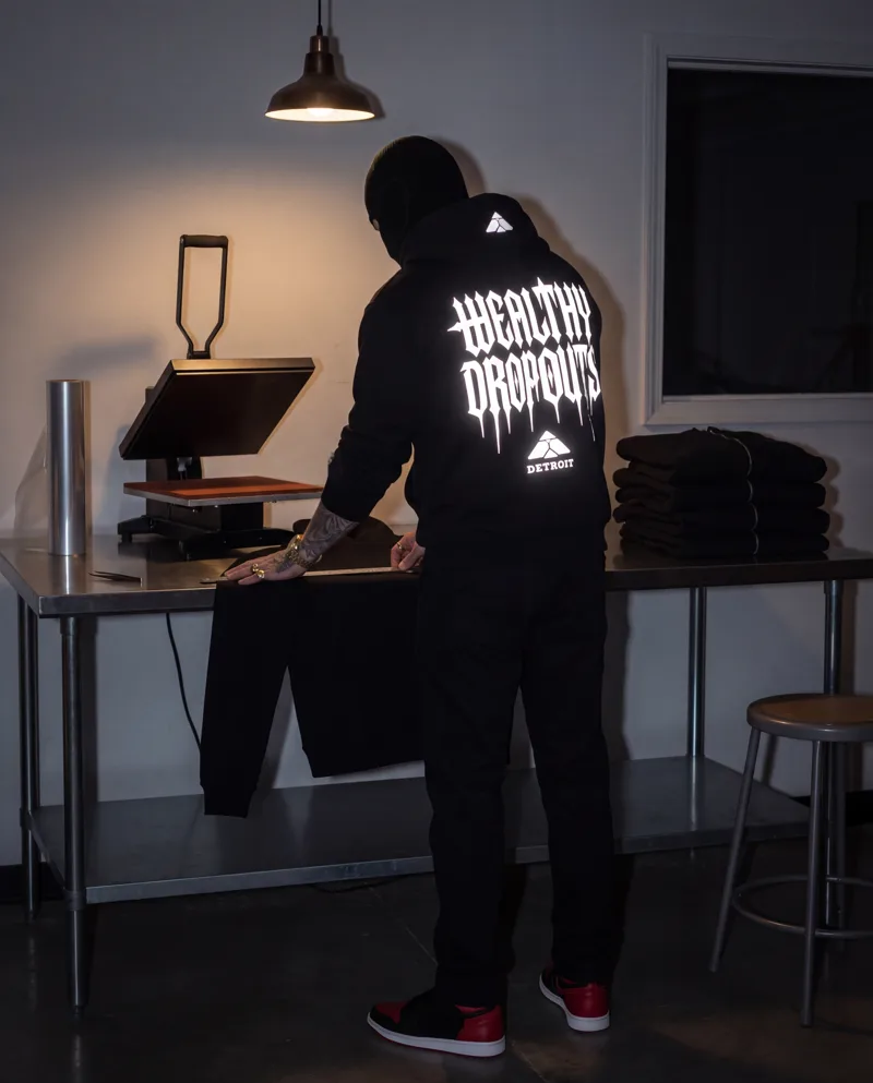 From behind: alone at a steel bench after hours beside a heat press, laying a ruler across a black hoodie, WEALTHY DROPOUTS lit white across his back.