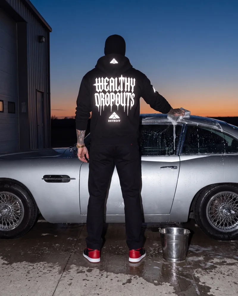 From behind: he washes a silver coupe himself at first light, the sponge on the roof, WEALTHY DROPOUTS lit white across his back.