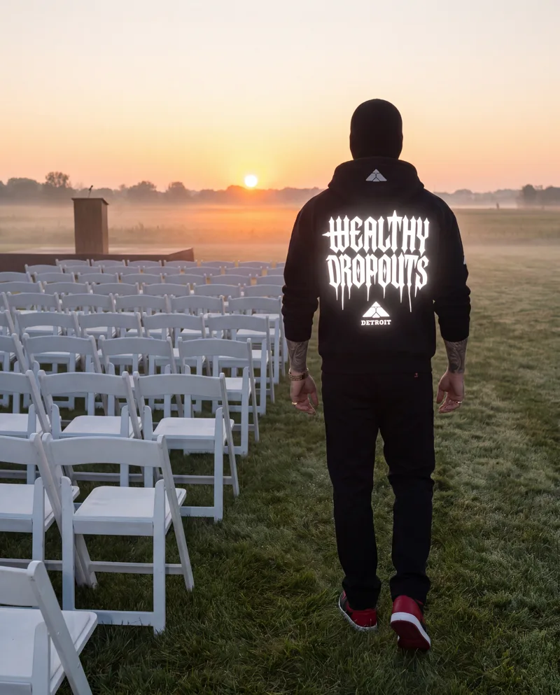 From behind: he walks past rows of empty white folding chairs toward the sunrise, WEALTHY DROPOUTS lit white across his back.
