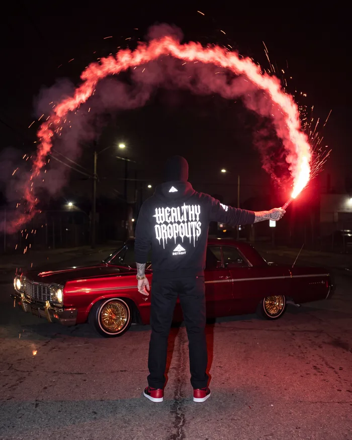 Worn, from behind: holding a red flare that has traced an arc across a night street
