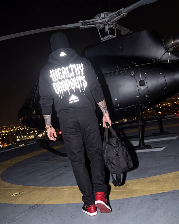 Worn, from behind: WEALTHY DROPOUTS in reflective gothic lettering across the back, the mark and DETROIT beneath, the mark on the hood; walking across a rooftop at night with a bag